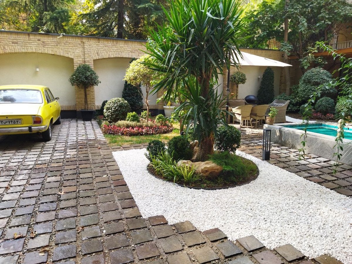 Courtyard with yellow car, tall tropical plant, brick patio, flowering garden beds, and swimming pool surrounded by ivy-covered walls