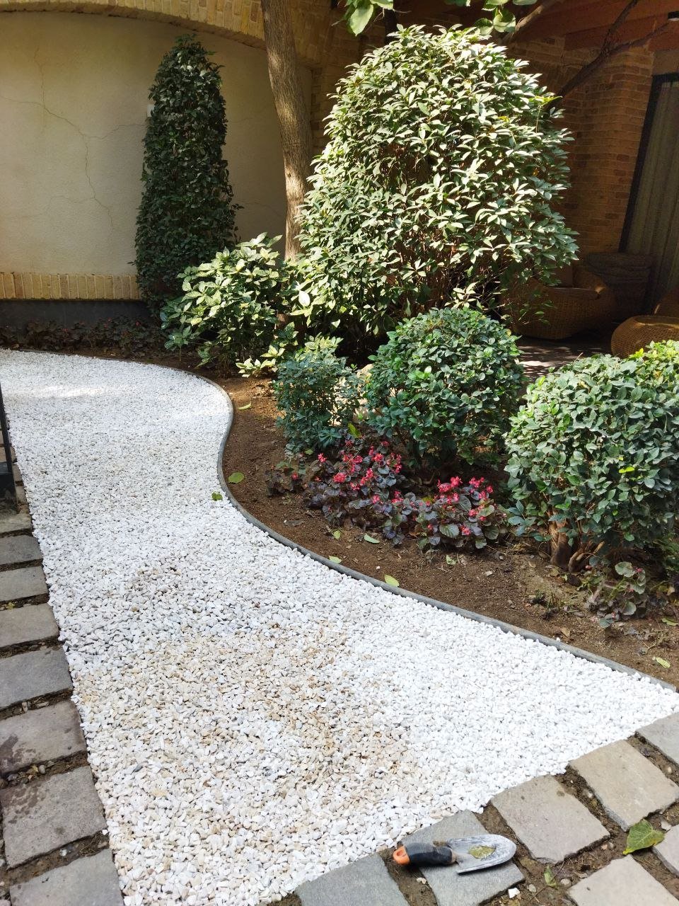 Curved gravel pathway through a garden courtyard with boxwood shrubs, flowering plants, and ivy-covered wall in background
