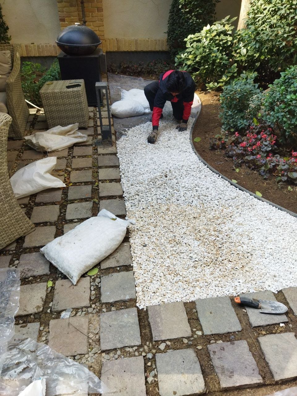 Person in black and red clothing installing white gravel in a backyard patio garden with paving stones and green hedges