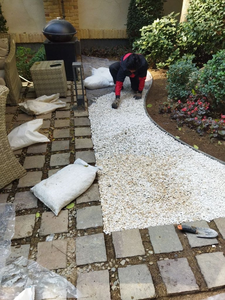 Person in black and red clothing installing white gravel in a backyard patio garden with paving stones and green hedges